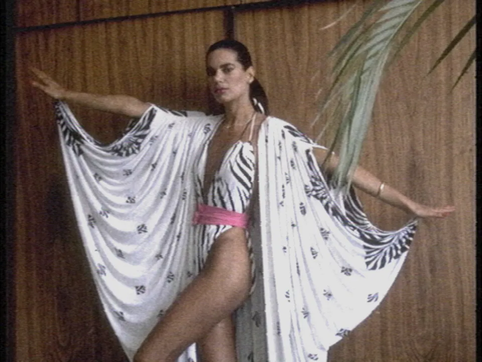 A 1980 swimwear model in a zebra-print swimsuit and a white cape, arms spread against wood panelling, clean apart from noise and a soft cast.